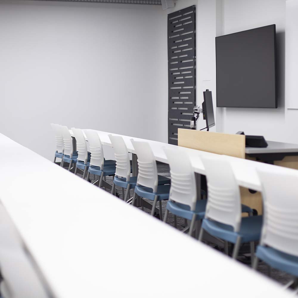 A modern, bright classroom featuring rows of white desks and blue-cushioned chairs facing a large flat-screen monitor and an instructor's podium