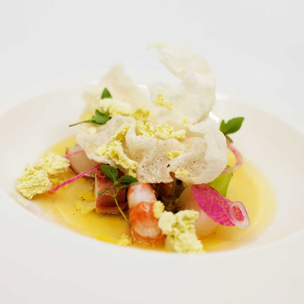 A delicate, foam-topped seafood dish featuring poached shrimp and crispy rice crackers.