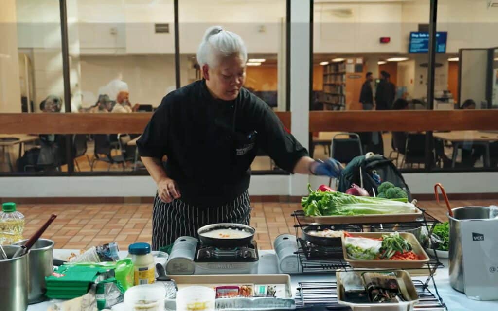 A chef prepares fresh vegetables and a simmered dish during a community cooking demonstration in an open indoor space