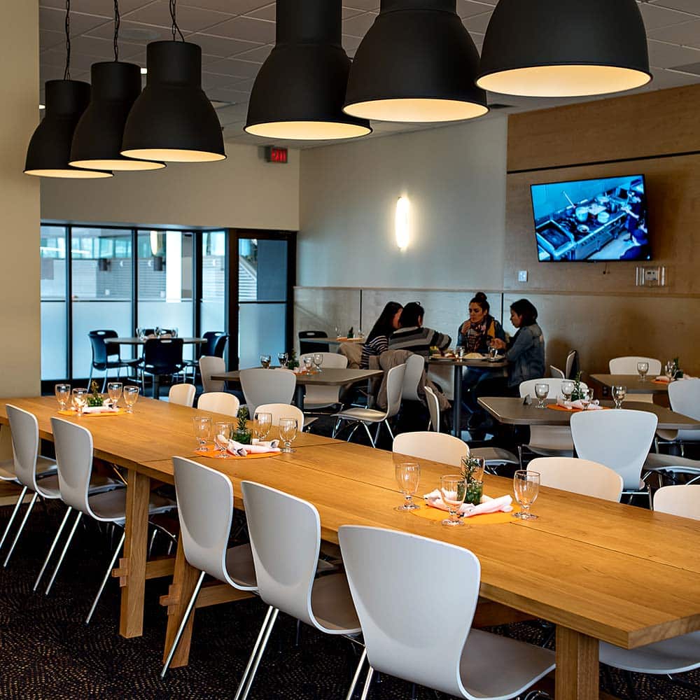 An inviting bistro dining area with long wooden communal tables, white modern chairs, and oversized black dome pendant lights.