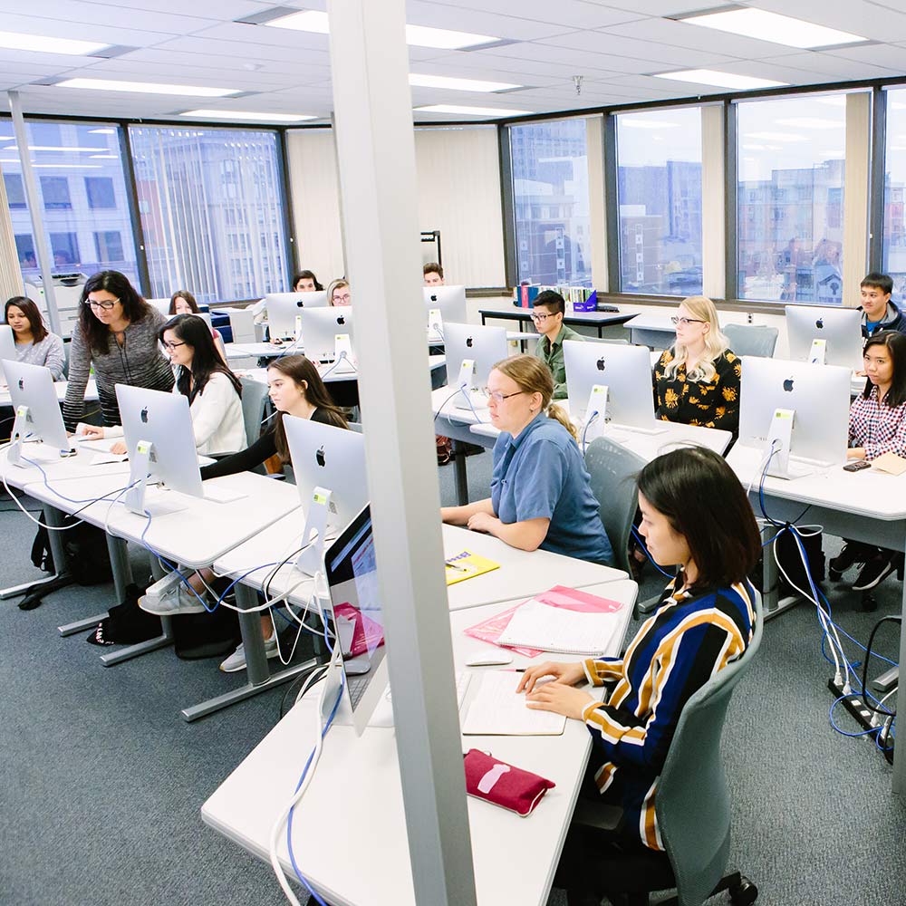 A group of students engaged in a lesson within a bright Mac computer lab equipped with rows of iMac workstations.