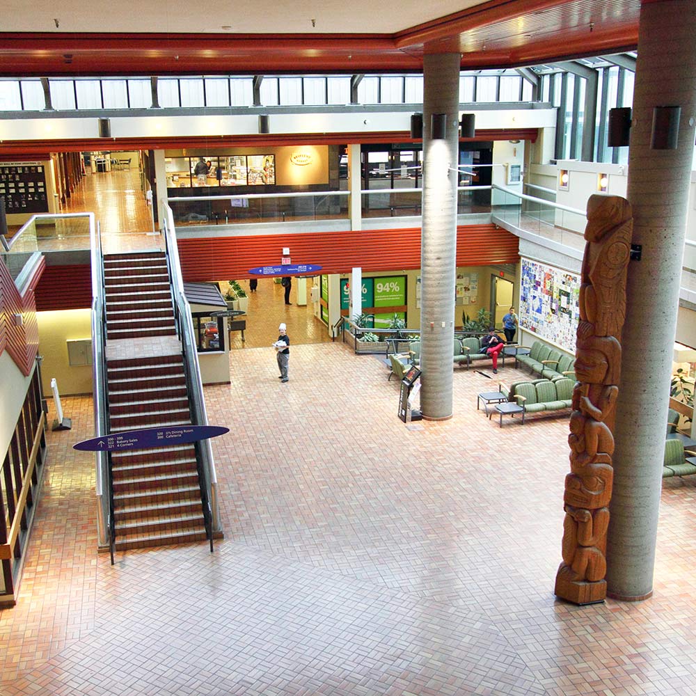 A high-ceilinged campus atrium with brick-patterned flooring, a grand staircase, and a large Indigenous totem pole standing near a seating area.