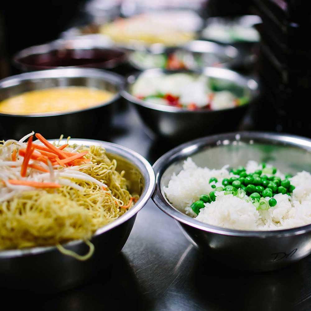 A variety of stainless steel prep bowls filled with noodles, rice with peas, and colorful stir-fry ingredients in a professional kitchen.