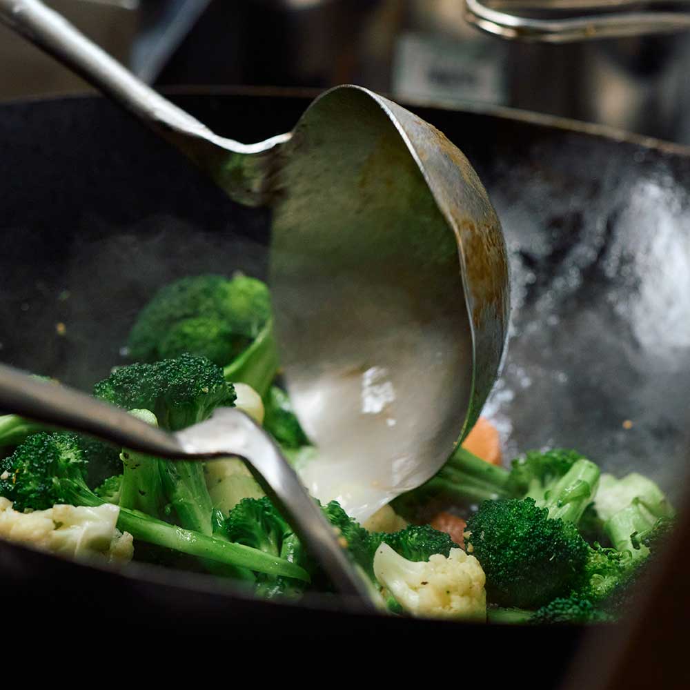 Fresh broccoli, cauliflower, and carrots being tossed with a metal ladle in a hot, steaming wok.