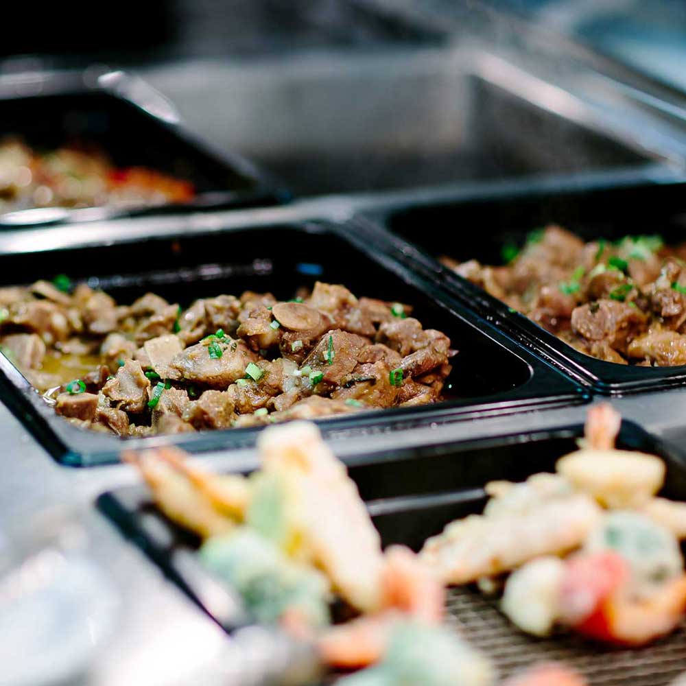 Close-up of various prepared Asian dishes including braised meats and tempura served in black buffet warming trays.