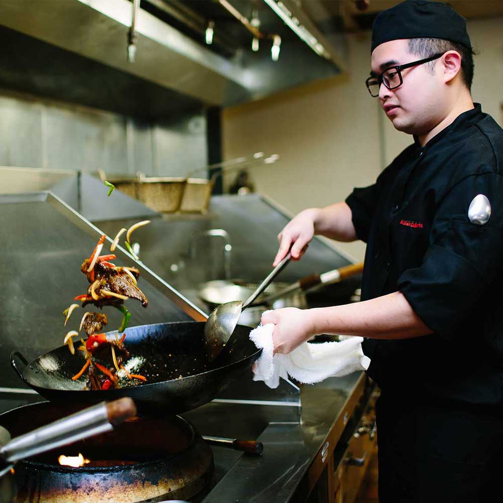 A professional chef in a black uniform skillfully tossing meat and vegetables in a large wok over a high-flame burner.