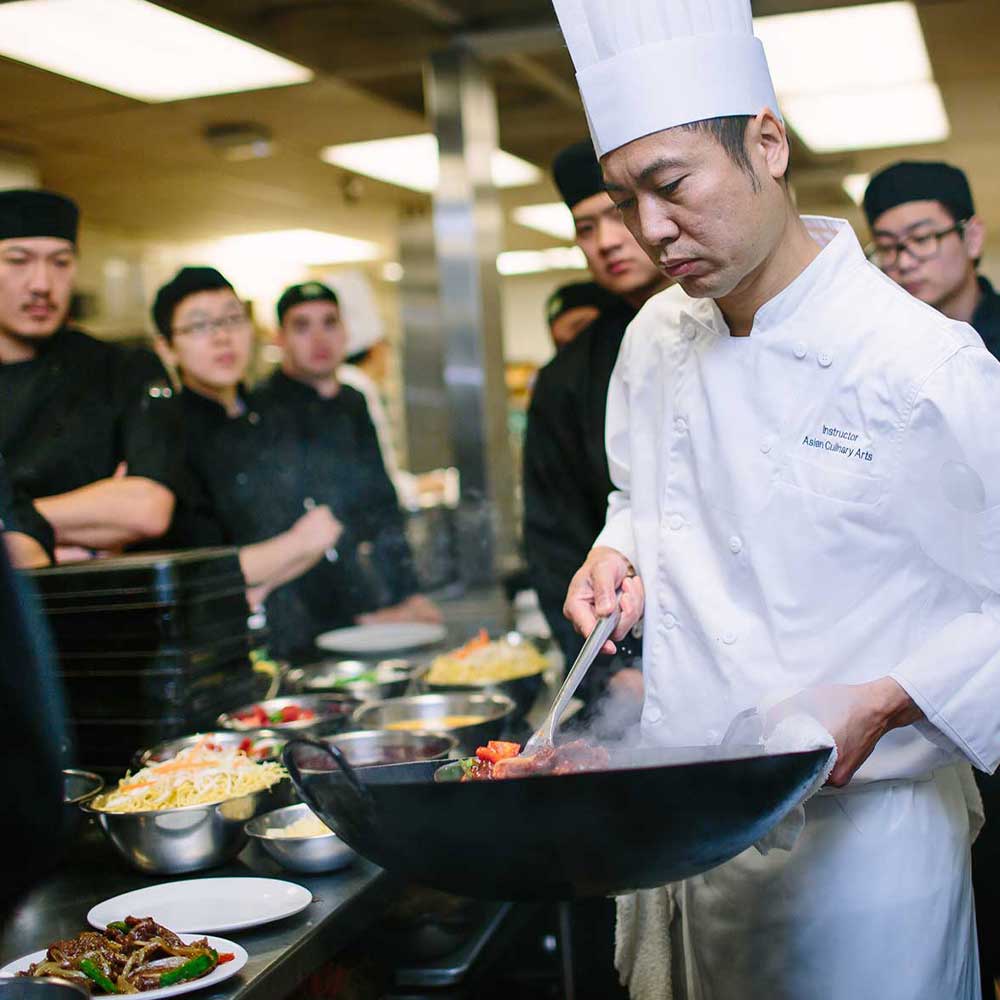 An Asian Culinary Arts instructor in a white chef's uniform demonstrates wok cooking techniques to a group of students.