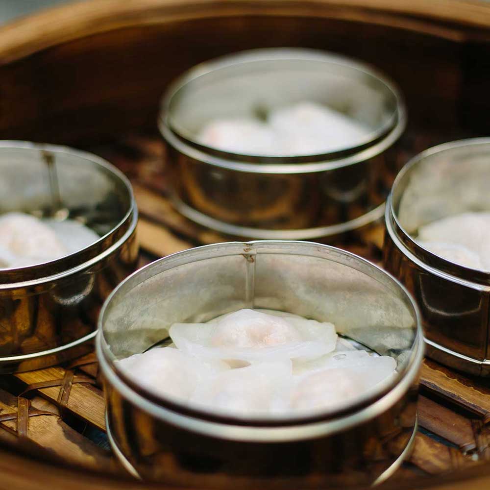 Four stainless steel steamer baskets containing delicate, translucent shrimp dumplings inside a traditional bamboo steamer.