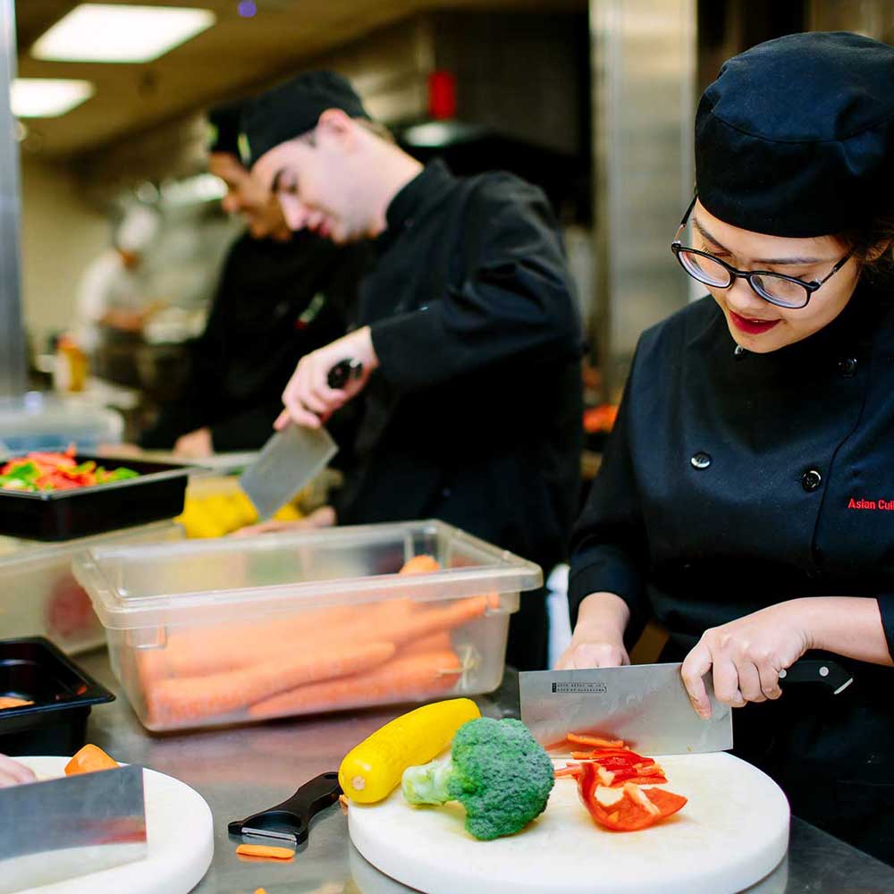 Culinary students in black uniforms focused on chopping fresh vegetables like peppers and broccoli on white cutting boards.