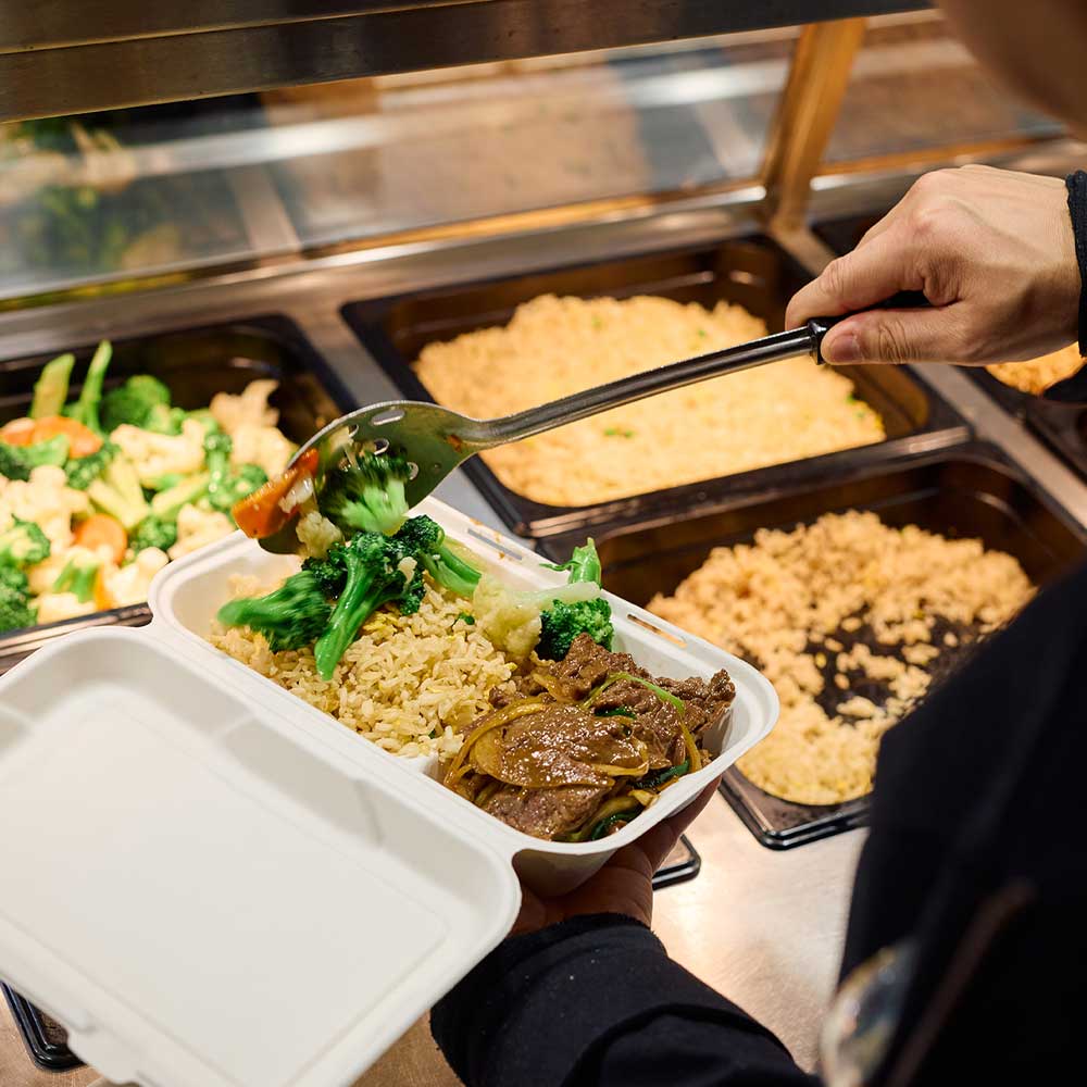 A person uses a serving spoon to fill a white compostable takeout container with fried rice, beef stir-fry, and steamed vegetables.