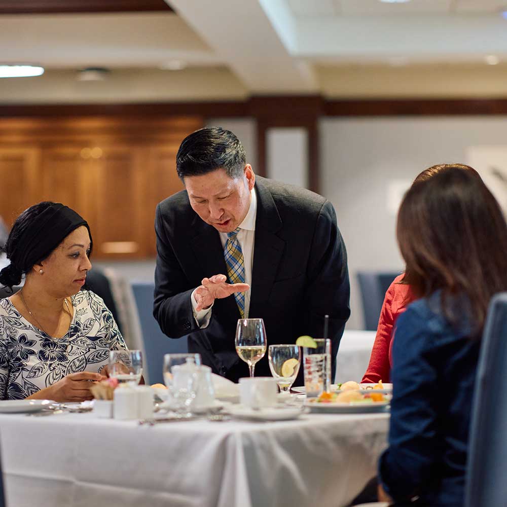 A professional host in a suit leans in to converse with guests seated at a white-clothed dining table.