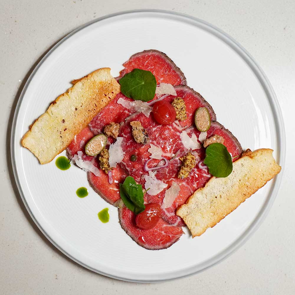 A refined plate of beef carpaccio topped with shaved parmesan, capers, and greens, served with toasted crostini.
