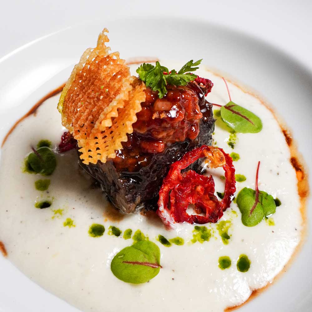 A tender braised short rib set upon a smooth potato purée, garnished with a crispy potato lattice and microgreens.