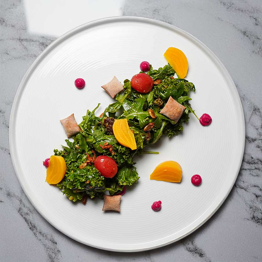 An artistic kale and arugula salad featuring golden beets, cherry tomatoes, and dollops of beet purée on a white plate.