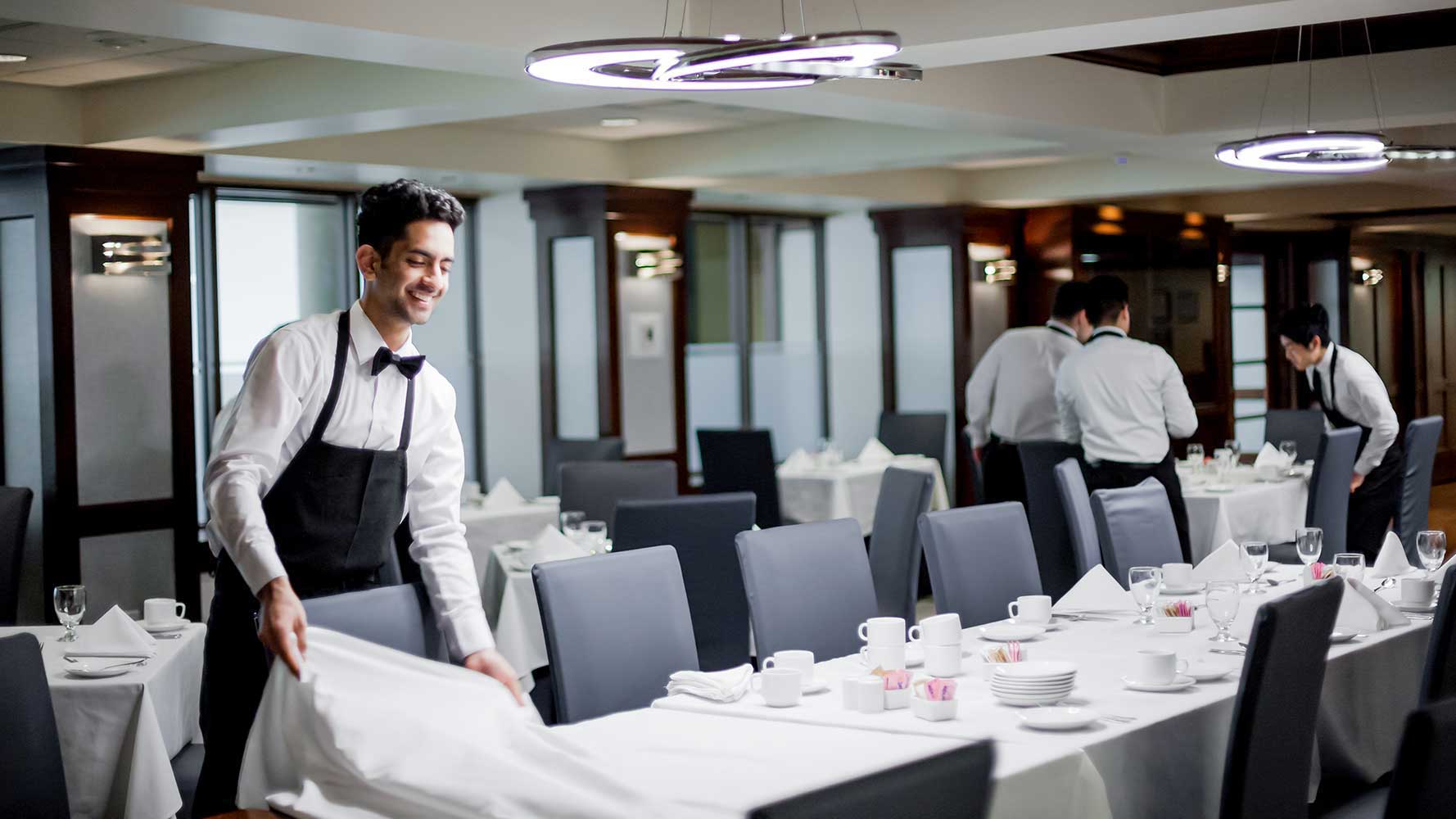 A smiling server in a formal bowtie and apron meticulously prepares a long dining table for a group event.