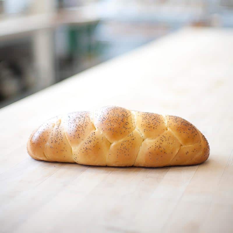 A golden braided loaf of bread topped with poppy seeds resting on a light wood countertop.