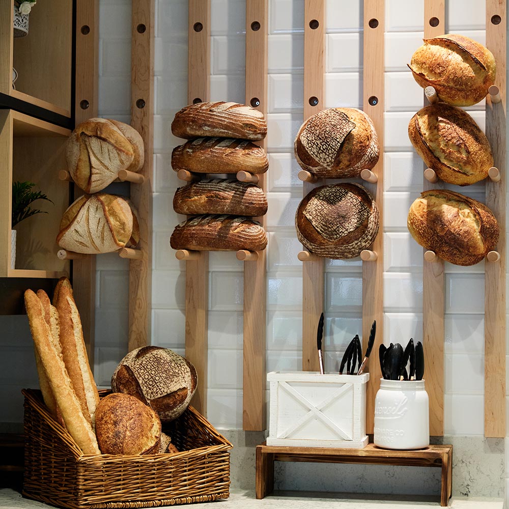An artisanal bread display featuring various sourdough loaves and baguettes on wooden wall pegs and in wicker baskets.