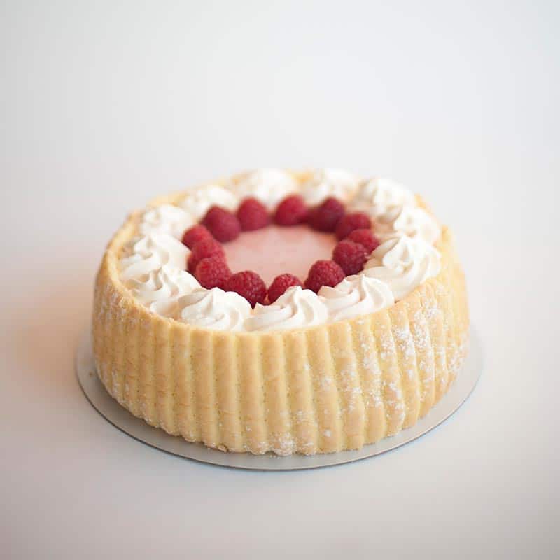 A round ladyfinger sponge cake topped with a ring of white whipped cream swirls and fresh red raspberries surrounding a pink glaze center.