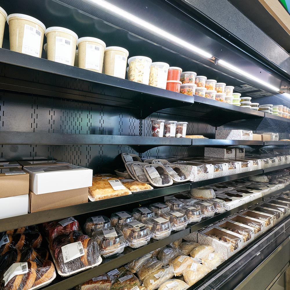 Fully stocked black refrigerated display shelves containing various packaged salads, sandwiches, donuts, sauces, and baked goods.