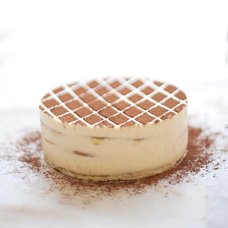 A round tiramisu cake featuring a diamond-patterned cocoa powder dusting on top, resting on a surface sprinkled with chocolate powder