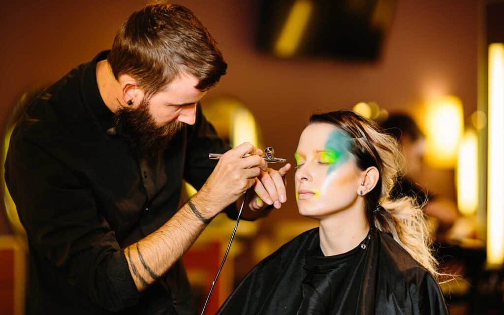 A makeup artist uses an airbrush tool to apply colorful green and blue makeup to a client.