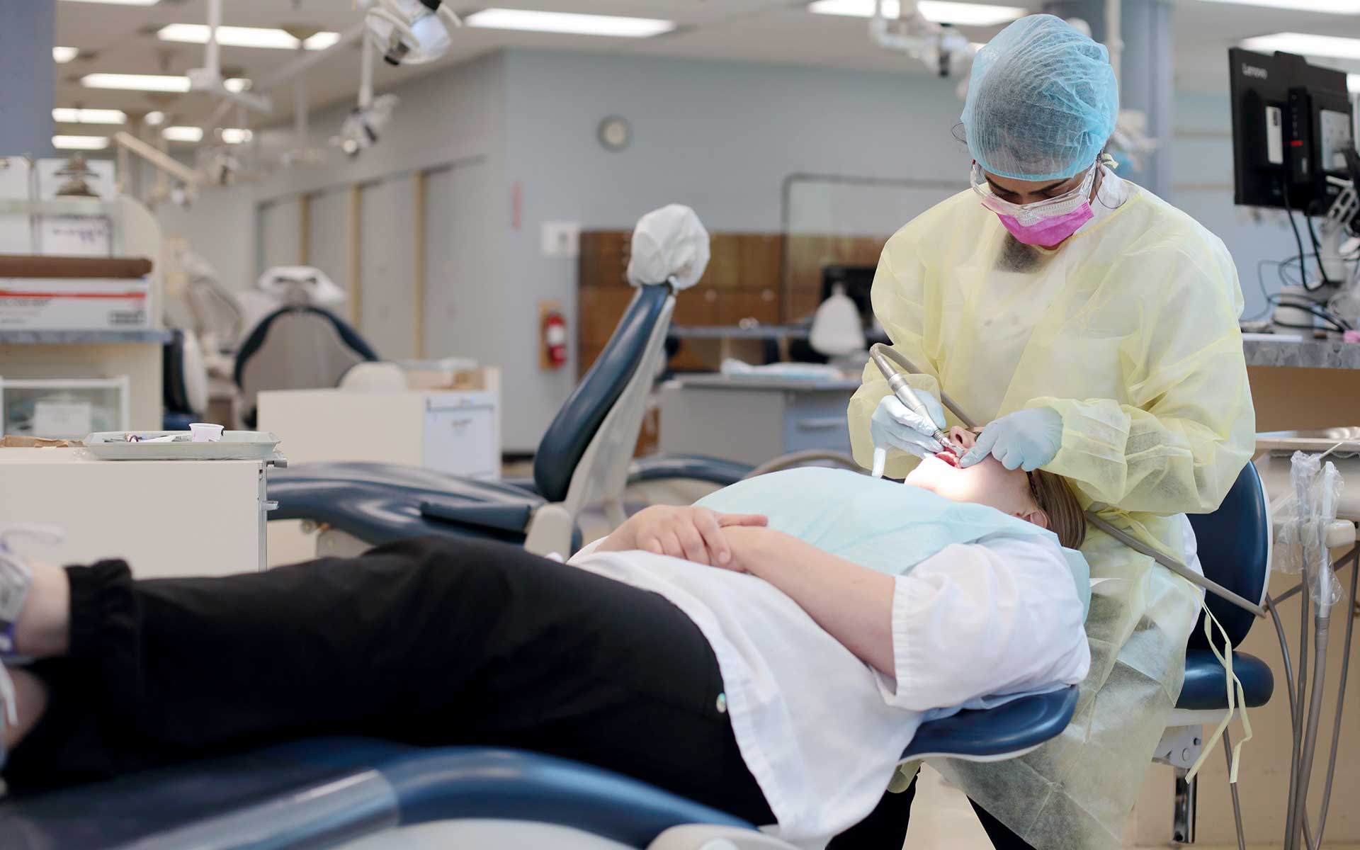 A dental student in protective gear performs a procedure on a patient in a clinic.
