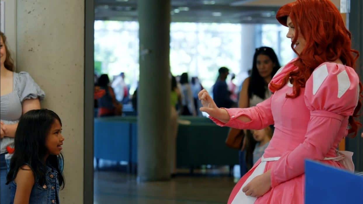 An entertainer dressed as a princess uses sign language to communicate with a young girl at an event.