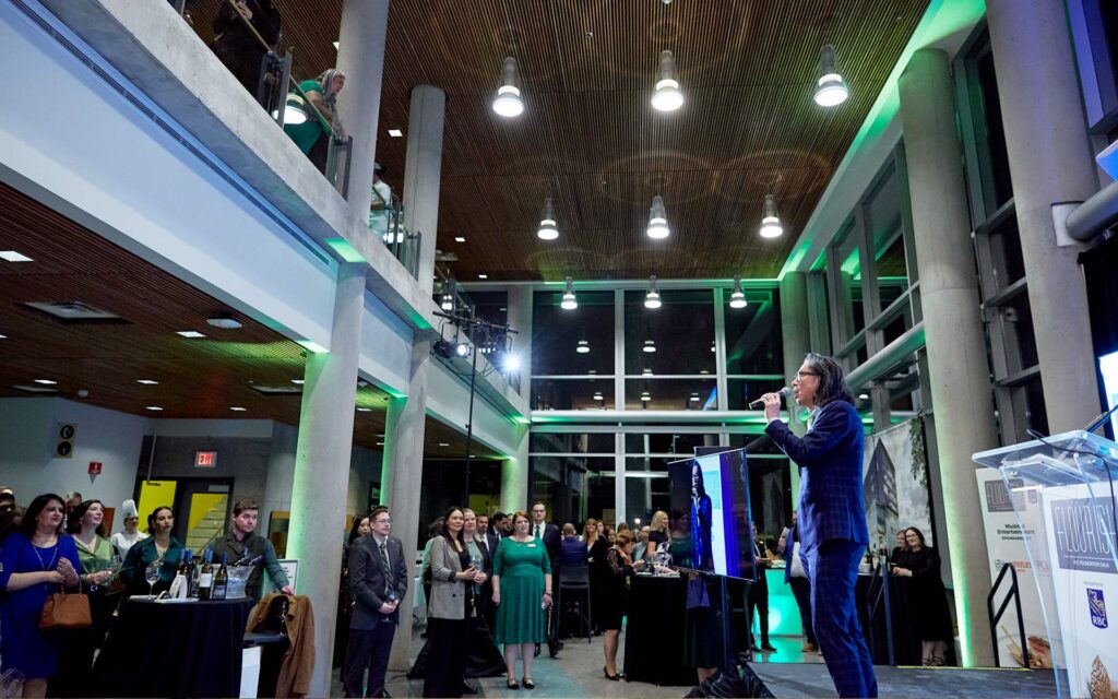 A man in a blue suit speaks into a microphone on a small stage during a formal gala in a modern, multi-level event space.