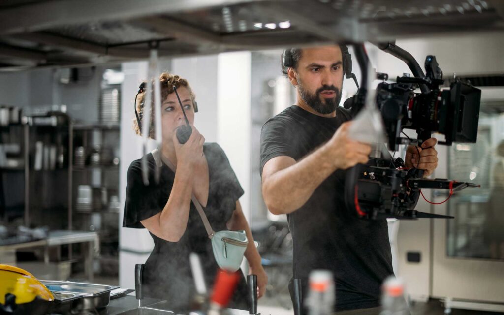 A man operates a professional film camera while a woman beside him speaks into a walkie-talkie on a movie set.