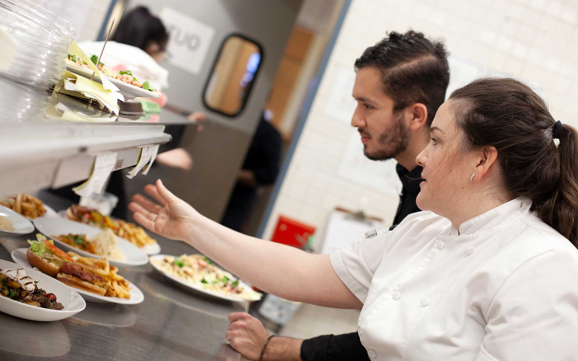 A chef and a kitchen staff member review order tickets at a busy service window filled with plated meals.