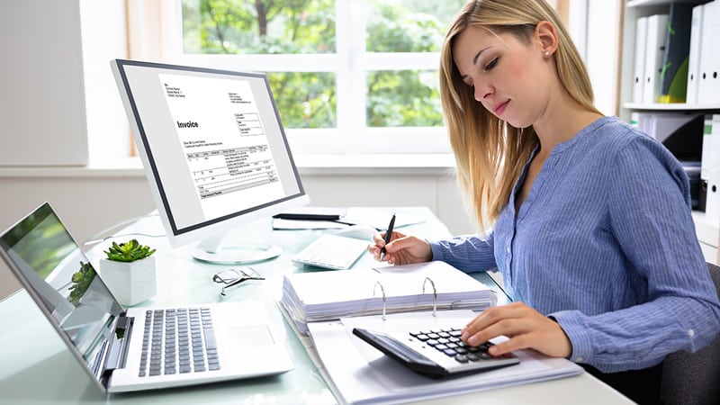 A woman works at a desk with a computer displaying an invoice. She uses a calculator and writes in a binder.
