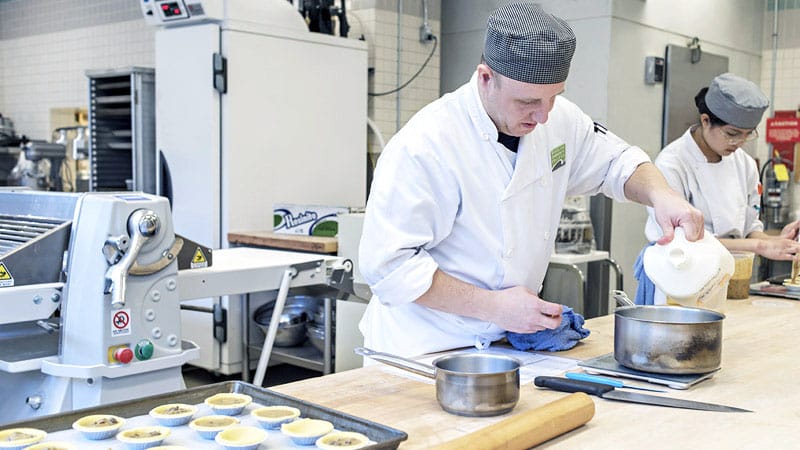 A chef in a white uniform and cap pours milk into a pot in a commercial kitchen. Another chef prepares dough nearby. The setting is organized and professional.