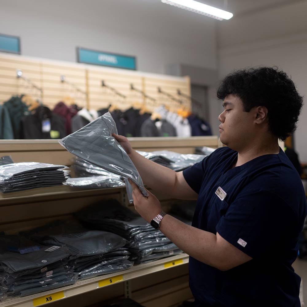 A student inspecting folded nursing uniform on the shelves of the well-stocked campus bookstore.