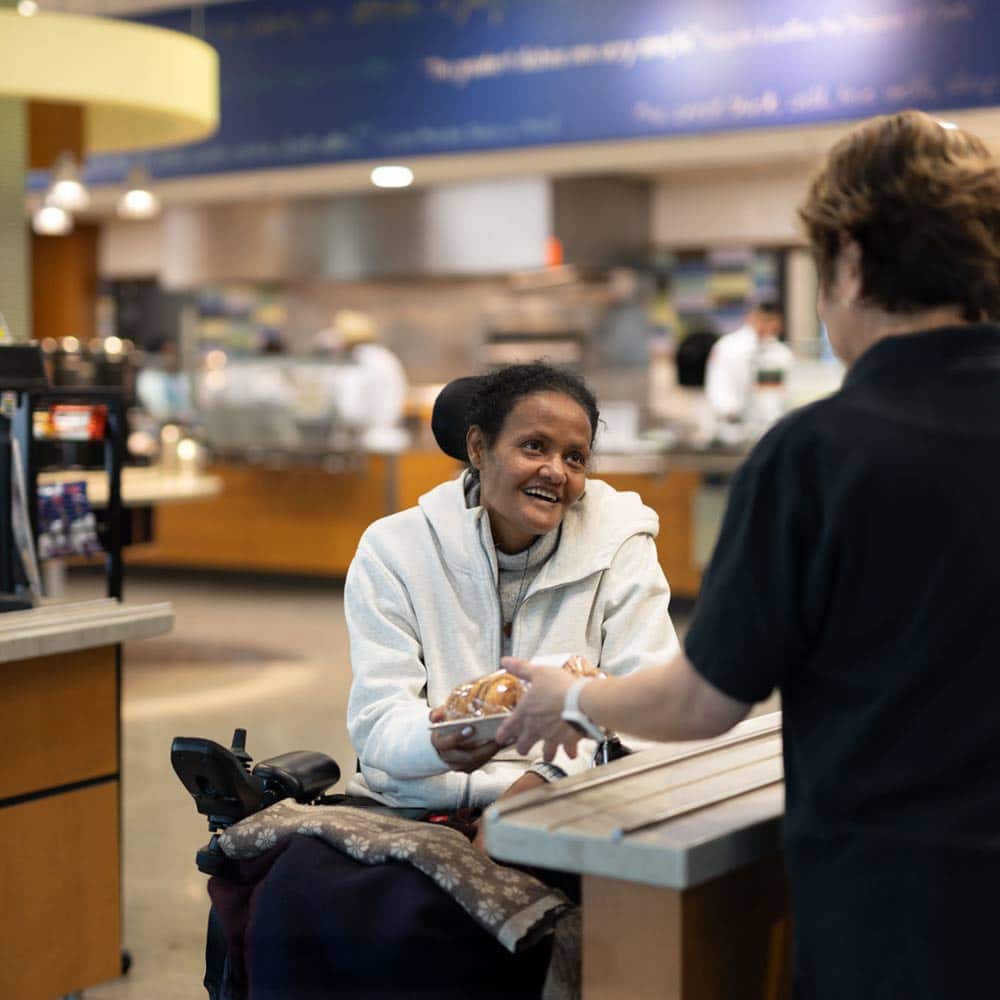 A friendly cafeteria staff member serves a student in a wheelchair, highlighting the campus's commitment to accessibility.