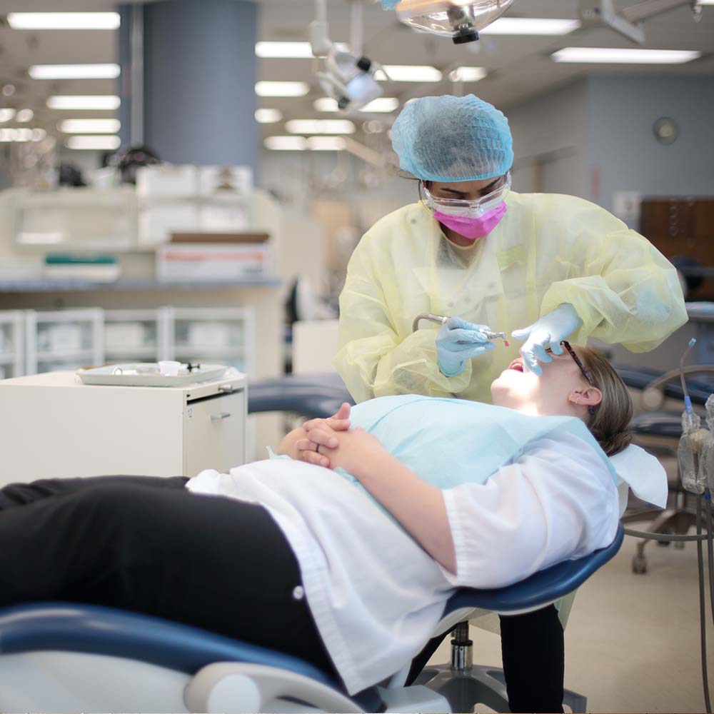 A dental student in full protective gear performs a procedure on a patient in a professional, brightly lit clinical training facility.