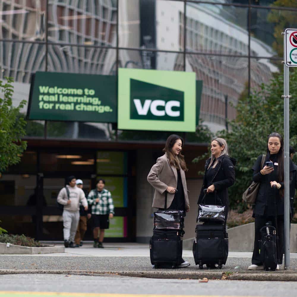 Students stand outside the VCC campus entrance near a bus stop, featuring a large green sign that reads "Welcome to real learning, for real change."
