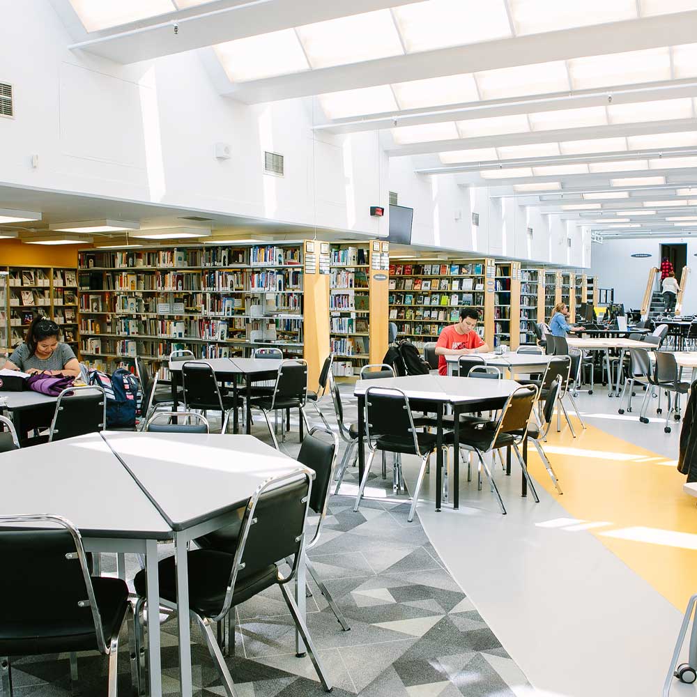 Students study at modular tables in a spacious, sunlit library filled with rows of books and large skylights.