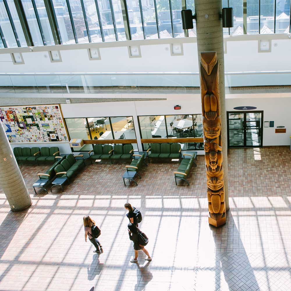n overhead view shows students walking through a large, airy atrium featuring high glass ceilings and a prominent carved totem pole.