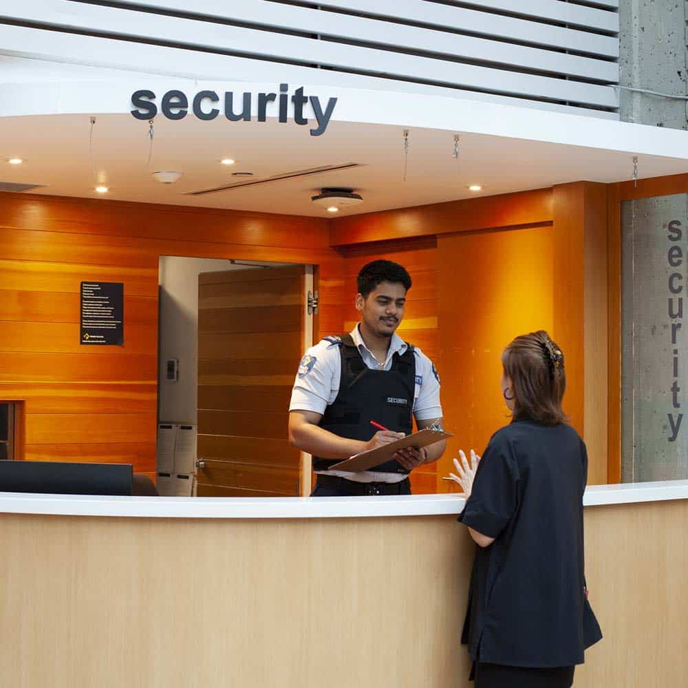 A uniformed security officer interacts with a student at a warm, wood-paneled service desk under a lit "security" sign.