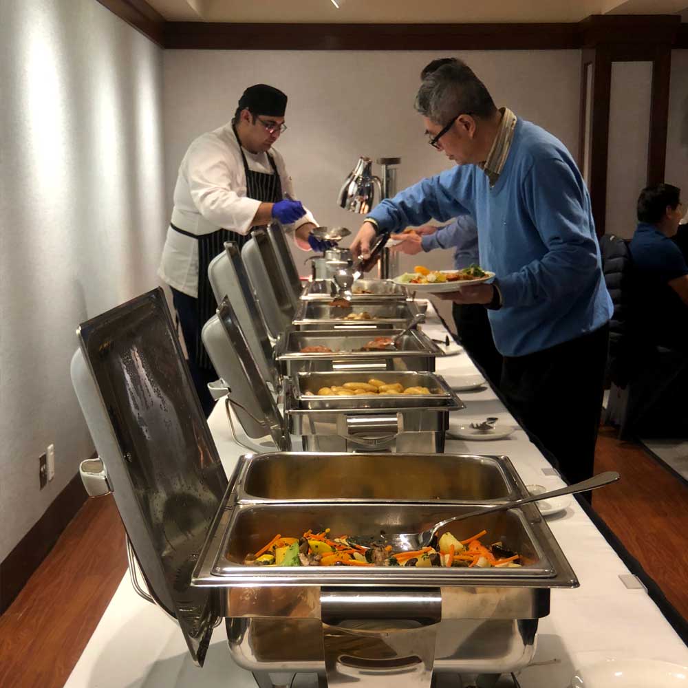 A chef in a white uniform assists a diner at a long buffet line featuring various hot dishes in silver chafing servers.