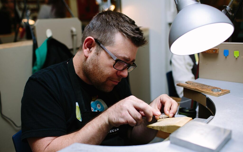 A focused jeweller wearing glasses works meticulously on a small piece of jewelry at a professional workbench under a bright desk lamp.
