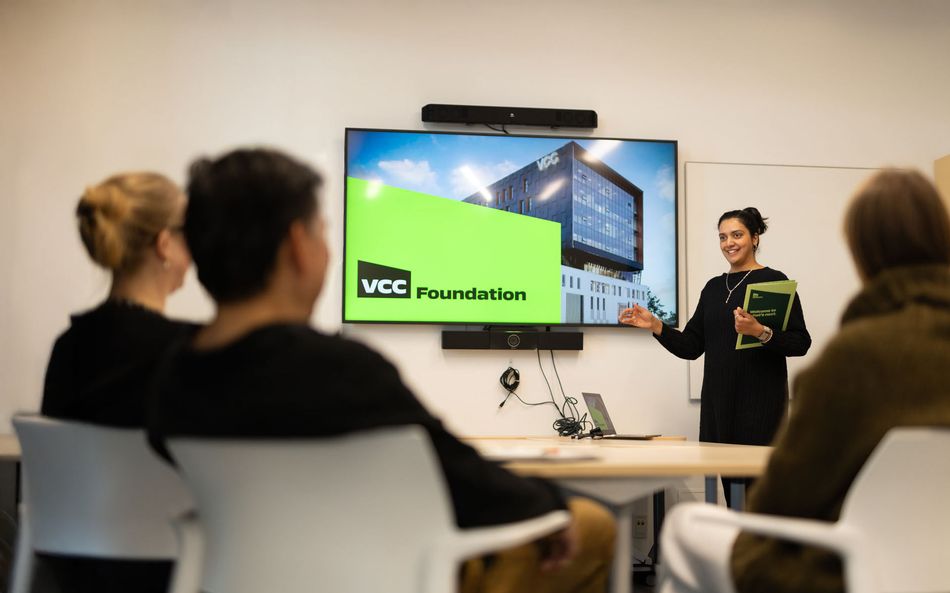A smiling presenter stands before a small audience, gesturing toward a screen displaying the VCC Foundation logo and a modern building exterior.