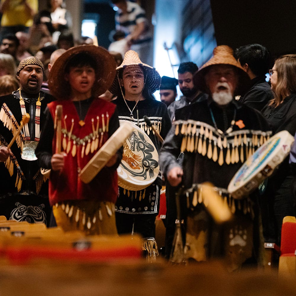 Indigenous performers dressed in traditional regalia are shown drumming and singing while processing through a crowded auditorium during a convocation ceremony.