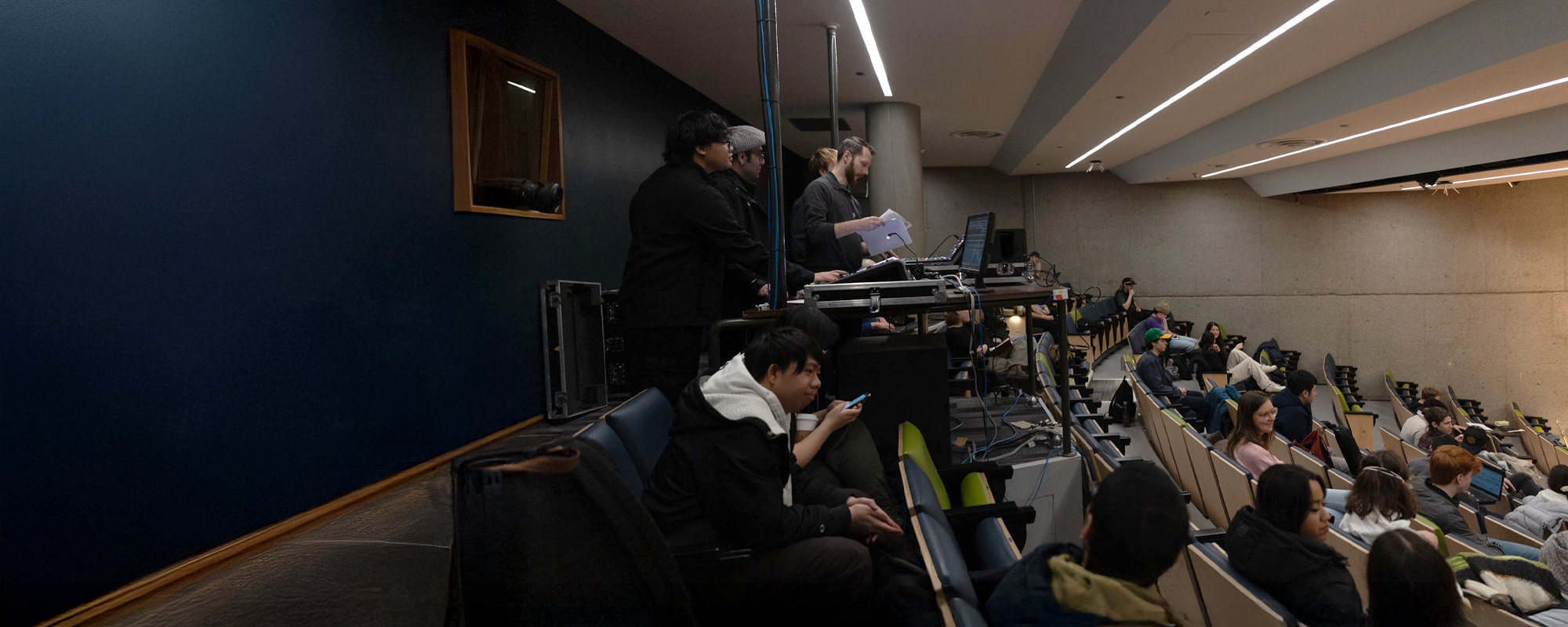 Students and staff are gathered in auditorium, where a technical crew manages AV equipment from a control booth.