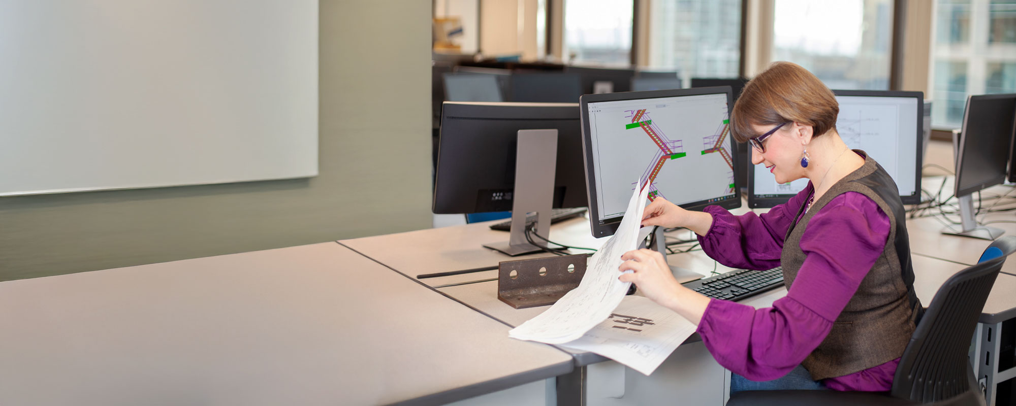A CAD and BIM student reviews printed building schematics while cross-referencing a 3D structural model on her computer monitor.
