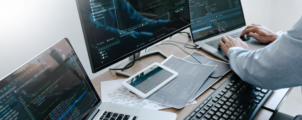 A programmer works at a multi-monitor desk setup, writing and reviewing code across several screens and a tablet surrounded by technical diagrams.