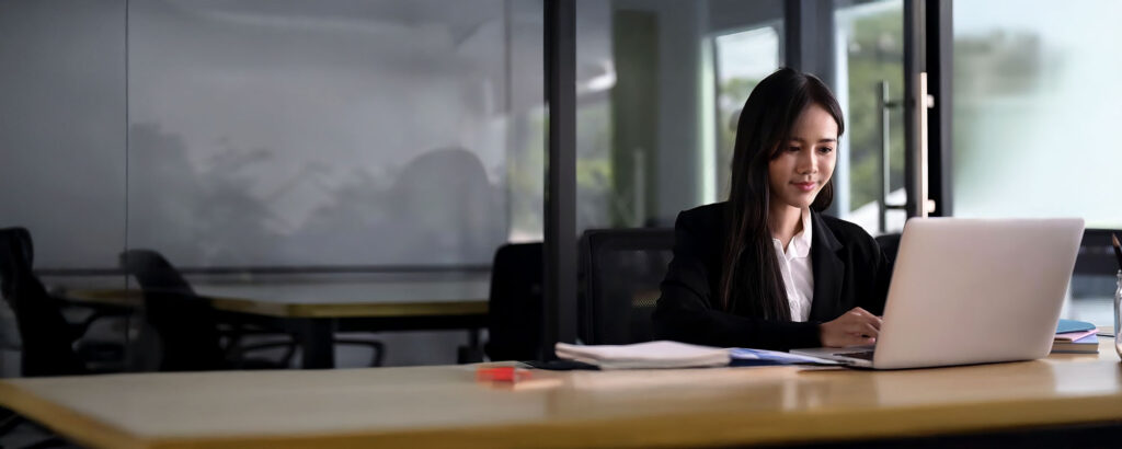 A business student in a blazer works diligently on her laptop at a desk in a modern, glass-walled office setting.