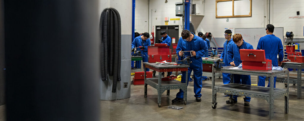 Several transportation trades students in blue coveralls work with hand tools at individual workbenches equipped with red toolboxes inside a spacious automotive training workshop.