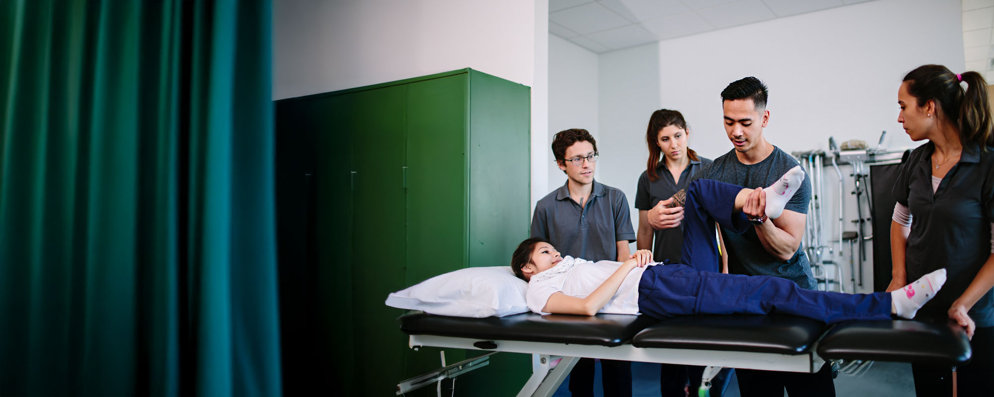 An instructor demonstrates physical therapy techniques on a patient lying on an examination table while a group of occupational and physical therapist assistant students observe.