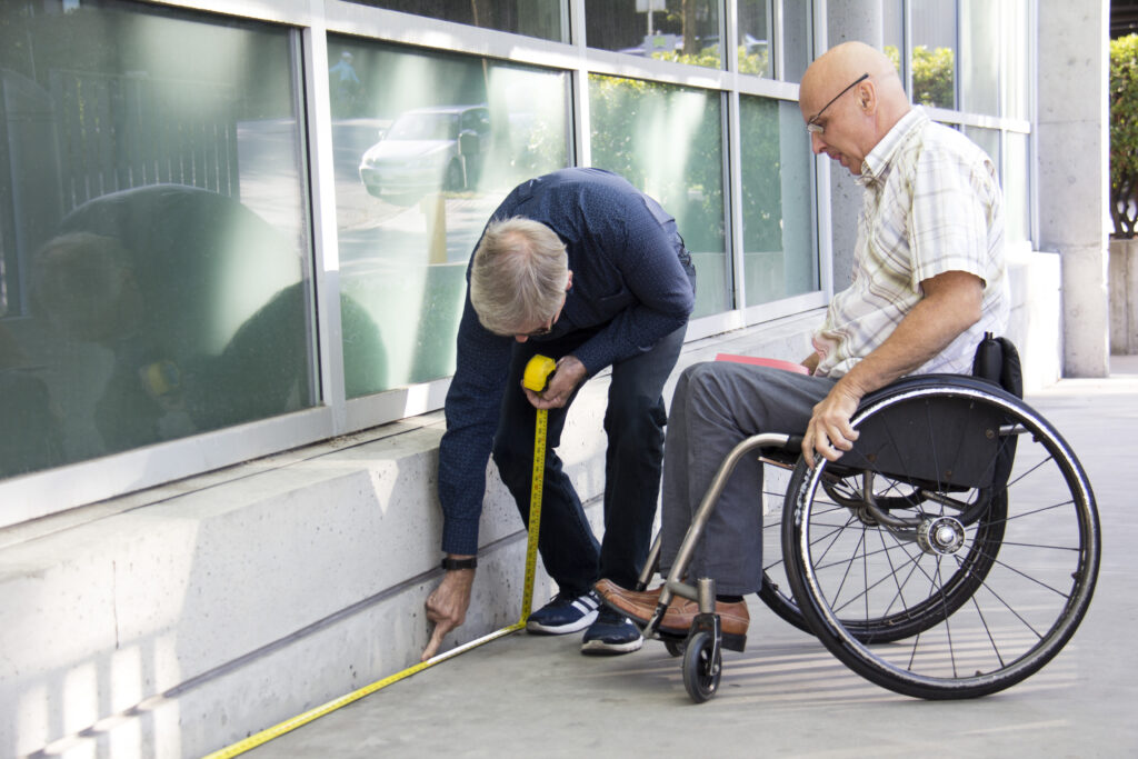 An accessibility assessment in progress, with a person measuring the entrance while a wheelchair user evaluates the space.